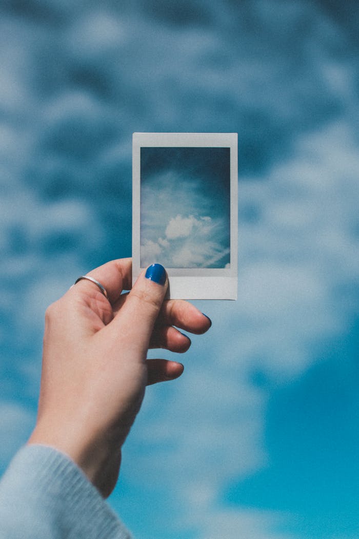 A hand with painted nails holds a Polaroid picture of clouds against a vibrant blue sky background.